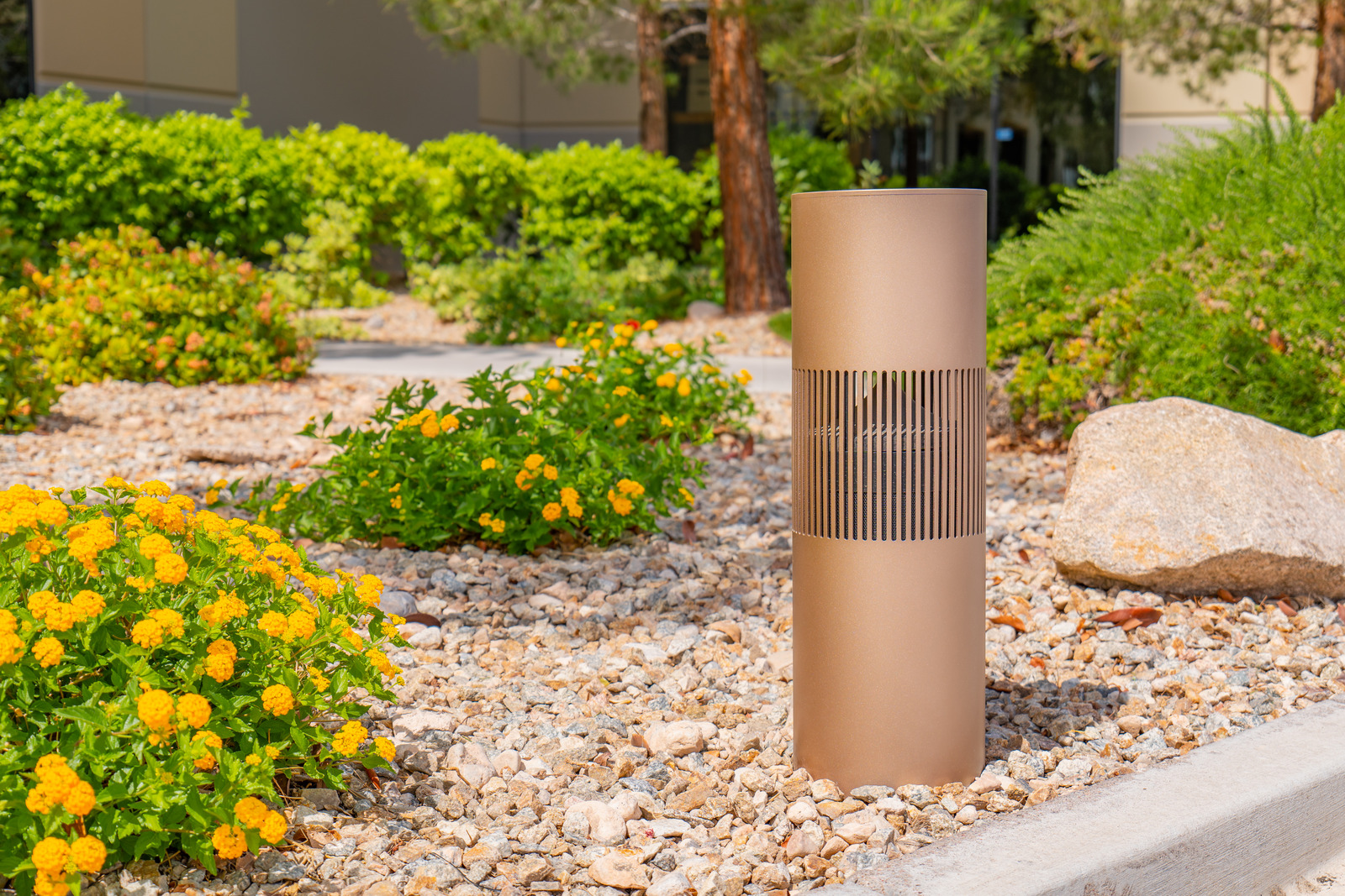 A bronze outdoor bollard speaker set into a gravel garden with flowering plants